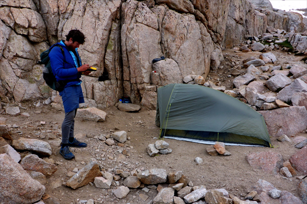 Man with unkempt, dark hair, wearing a blue jacket and blue shorts over tights looks down at object in his hands, as he stands near a tent in an arid rocky area.