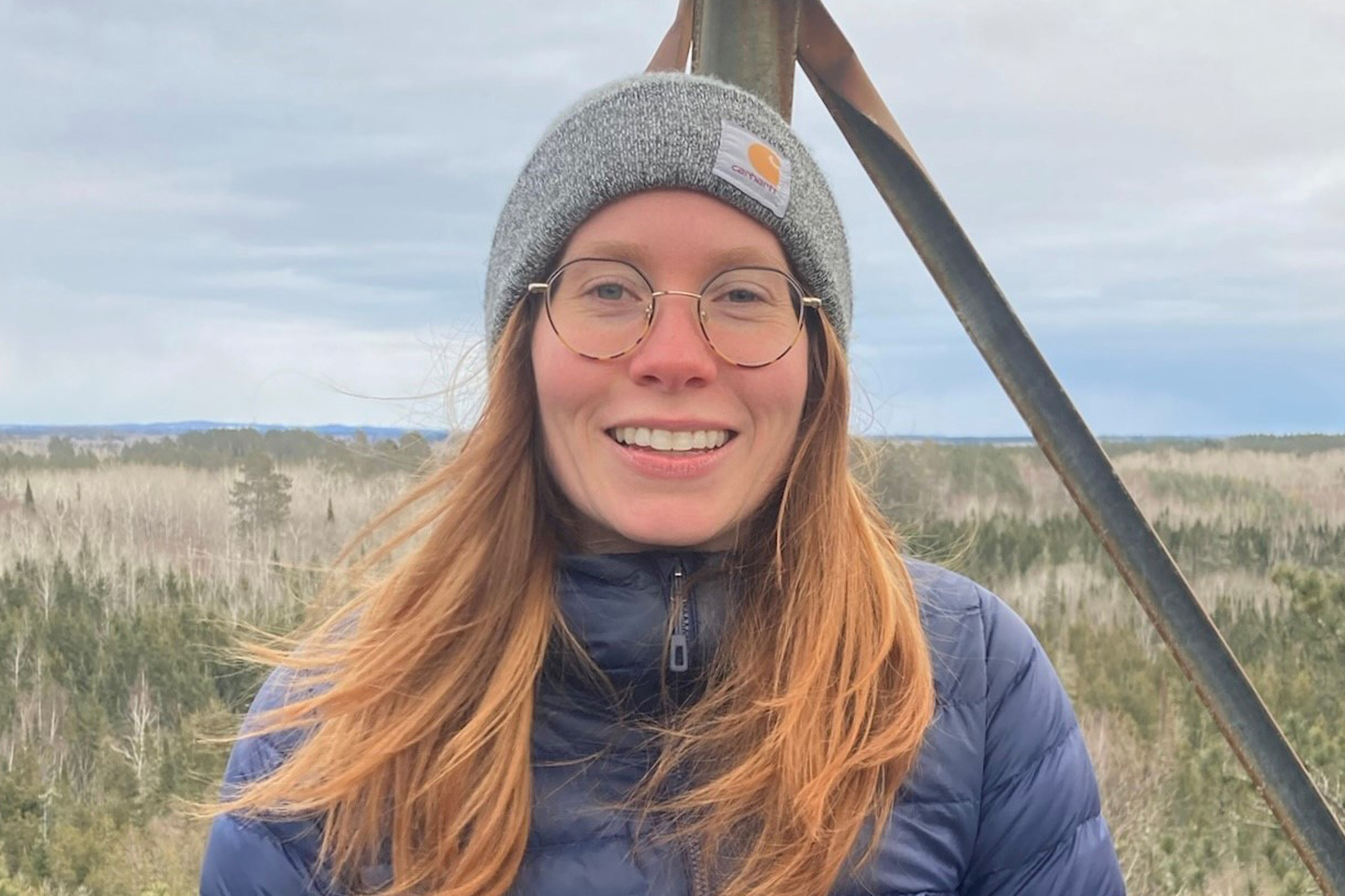 Woman in a stocking cap and glasses smiles at the camera while standing in an expansive prairie environment.