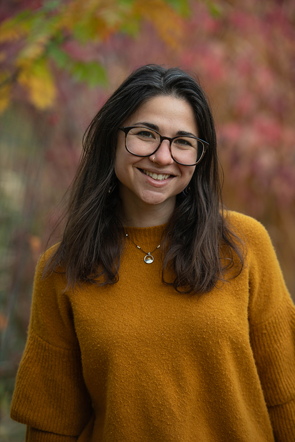 Woman wearing yellow sweater and glasses smiles, surrounded by autumn vegetation.