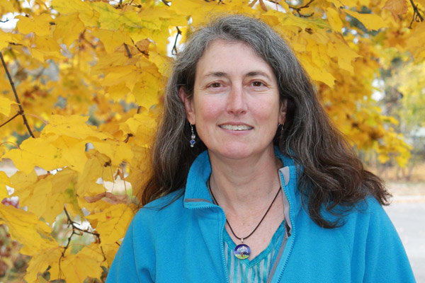 Woman with long, graying hair, wearing a blue shirt and jacket, smiles at the camera in front of a background of bright yellow maple leaves.